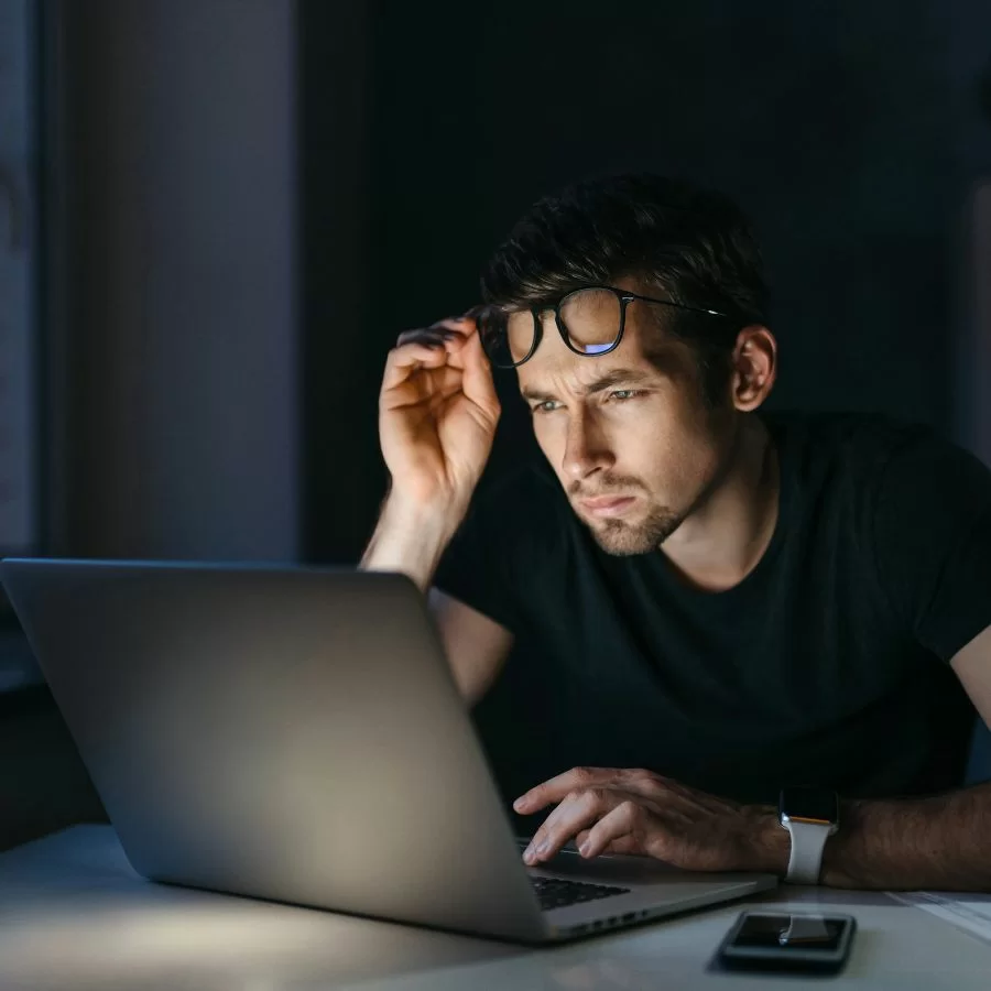 Người đang ông nheo mắt, đẩy kính lên khi nhìn chằm chằm vào laptop trong bóng tối, thể hiện sự mệt mỏi